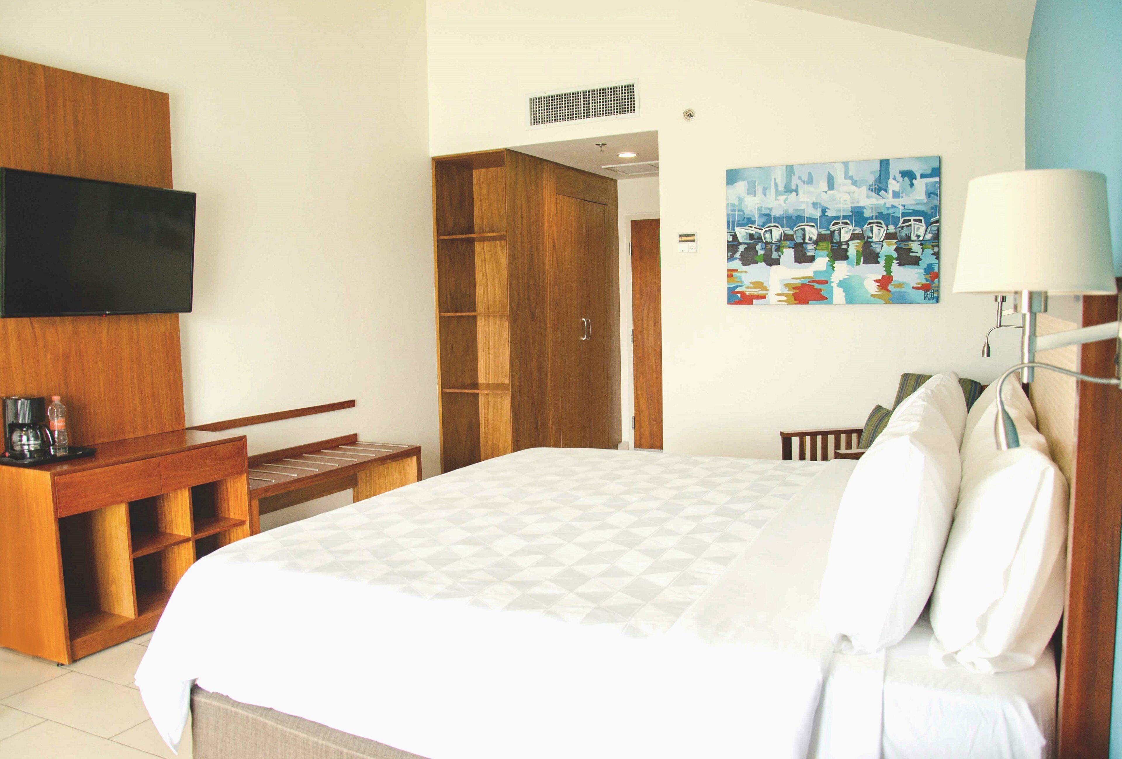 Een hotelkamer met een bed, een aan de muur bevestigde televisie, een schilderij en een bureau met een koffiezetapparaat.