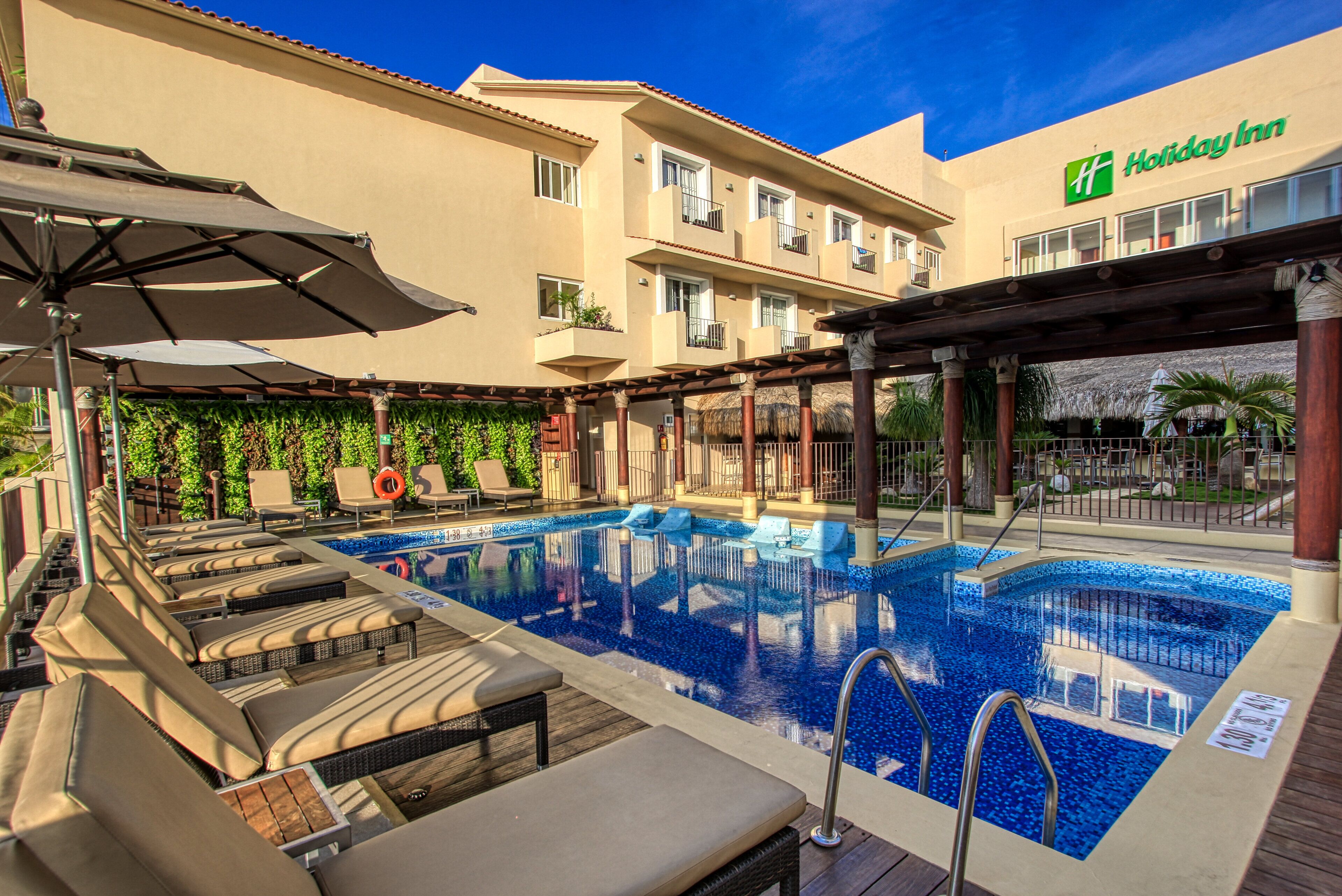 Zona de piscina de hotel con sillones de descanso y sombrillas, una piscina azul cristalina y un edificio con letrero Holiday Inn.