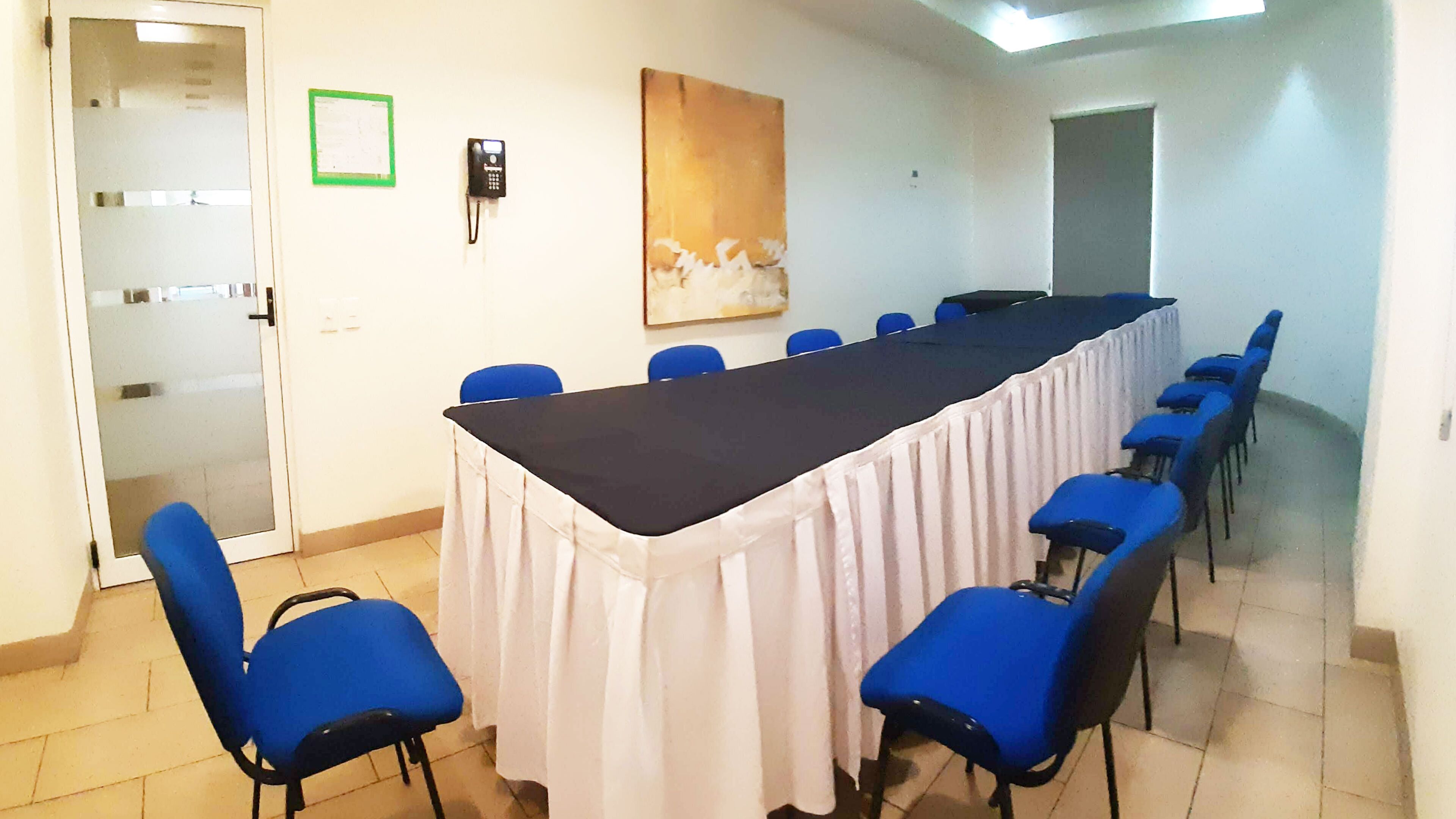 A conference room with a long table, blue chairs, a white curtain, a painting on the wall, and a door with a green sign.