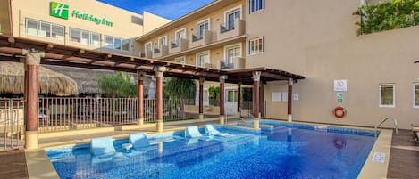 A hotel pool area with a clear blue pool, wooden deck, and a covered seating area.