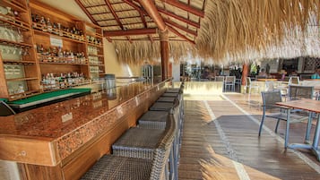 A bar area with a thatched roof, wooden shelves with bottles, and wicker seating.