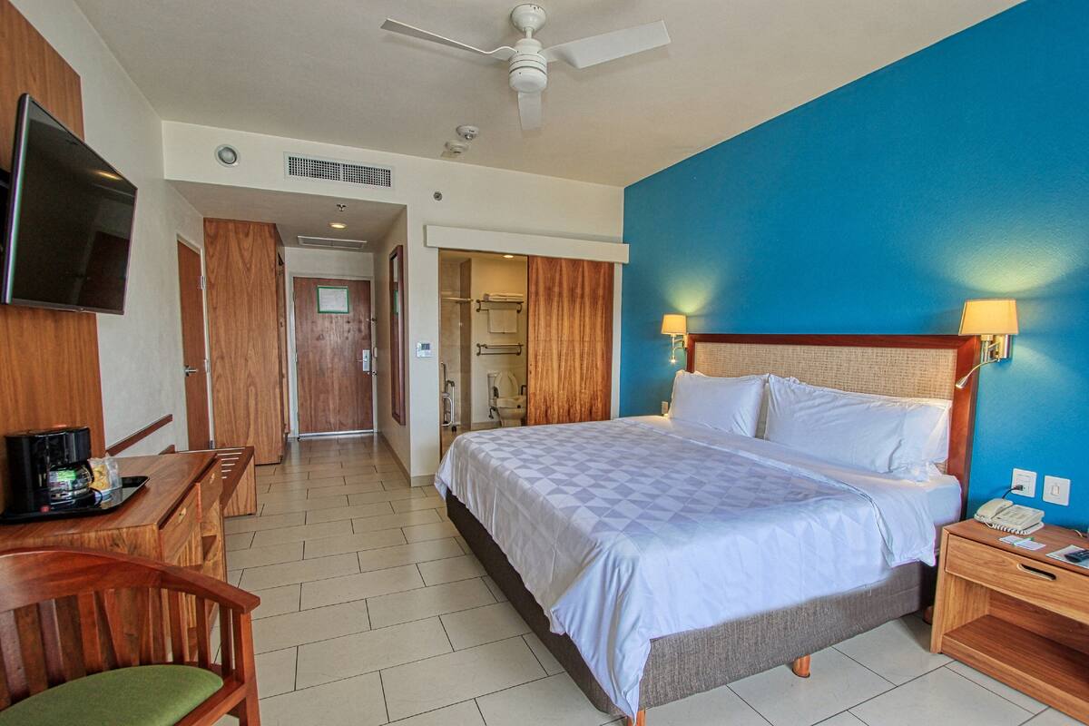 A hotel room with a bed, a desk with a chair, a television, and a bathroom visible through an open door.