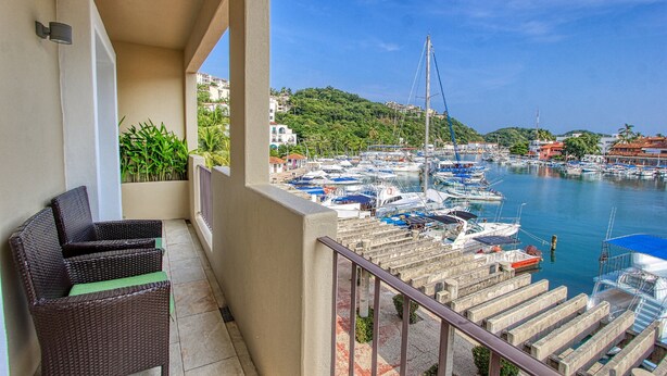 Un balcon agrémenté de meubles en osier, donnant sur une marina où sont amarrés des bateaux, avec une chaîne de collines en arrière-plan.