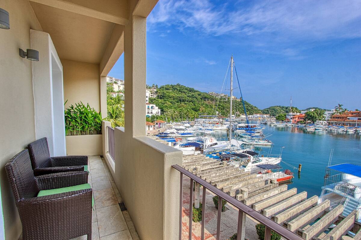 A balcony with wicker furniture, overlooking a marina with boats and a hilly backdrop.