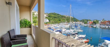 A balcony with wicker furniture, overlooking a marina with boats and a hilly backdrop.
