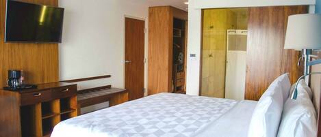 A hotel room with a bed, a desk with a chair, a television mounted on the wall, and a wardrobe.