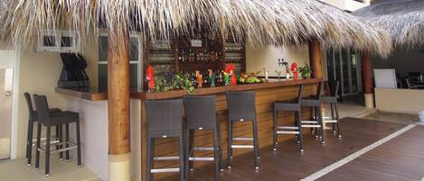 Uma área de bar com telhado de palha, balcão de madeira e cadeiras de vime.