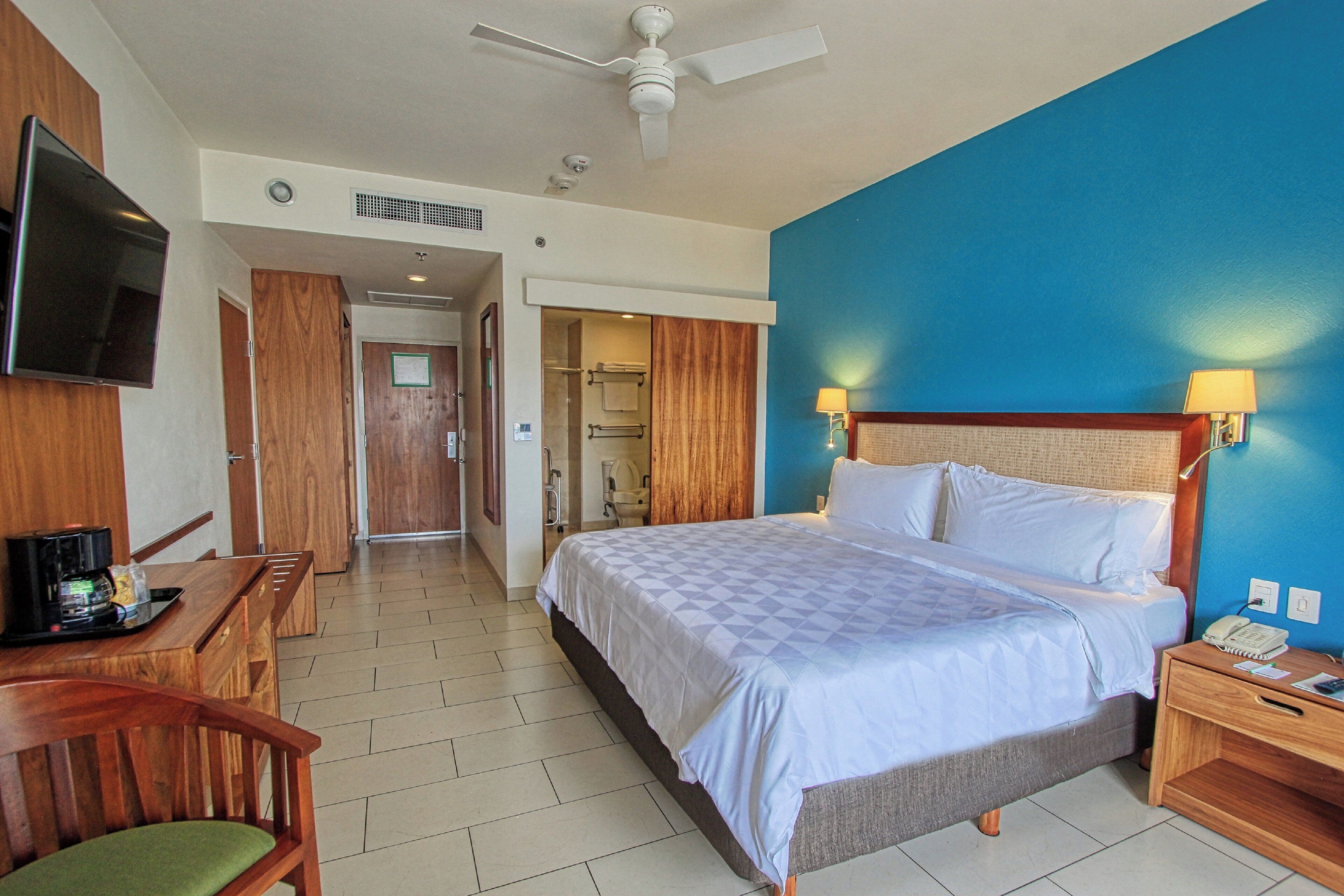 A hotel room with a bed, a desk with a chair, a television, a nightstand with a lamp, and a bathroom visible through an open door.
