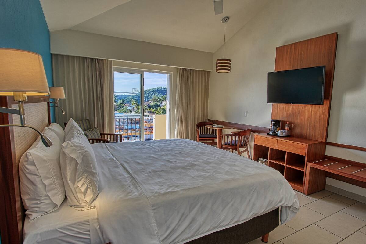 A hotel room with a large bed, a desk with a chair, a TV mounted on the wall, and a view of the outdoors.