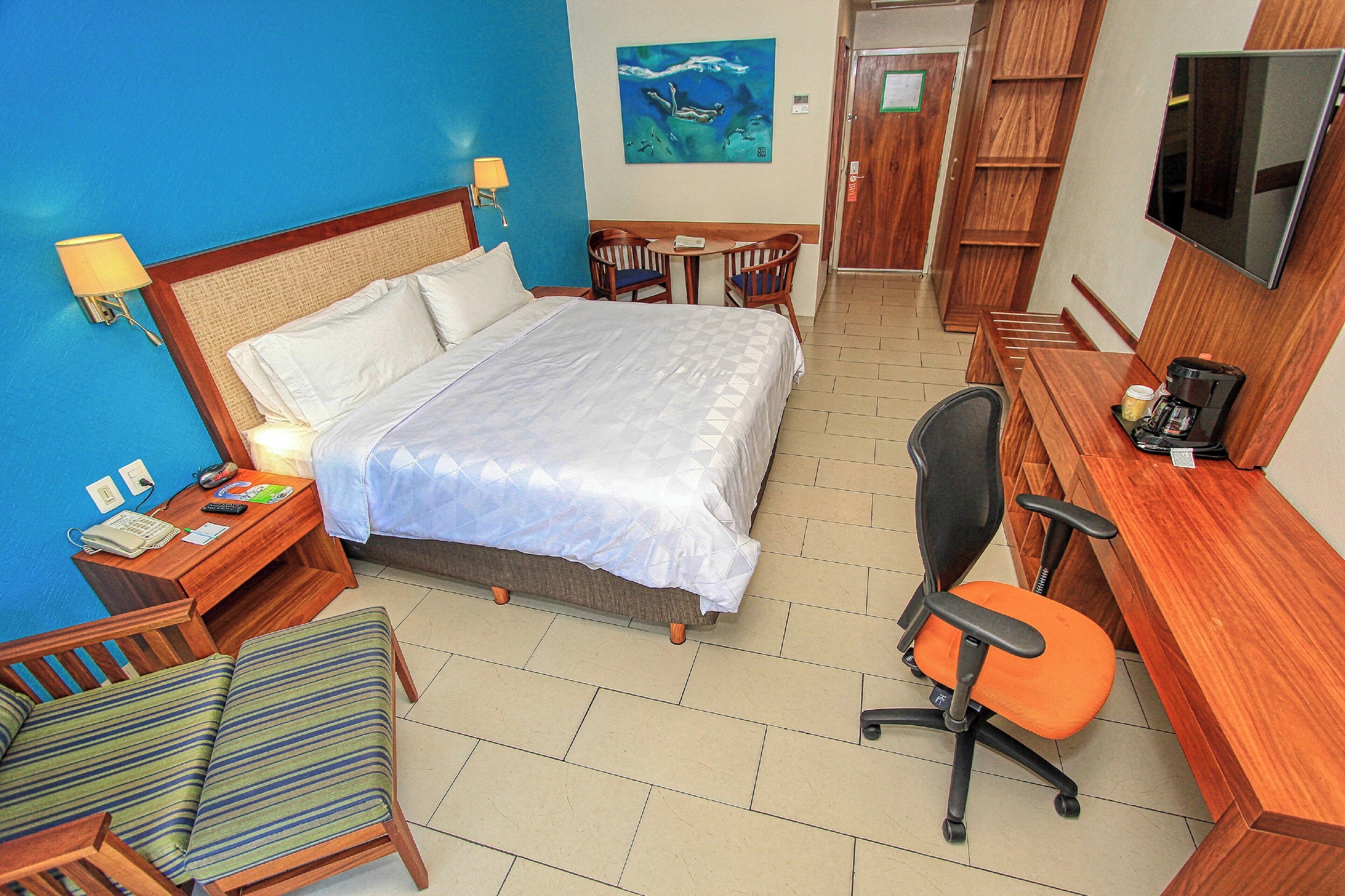 A hotel room with a bed, a desk, a chair, a TV, and a painting on the wall.