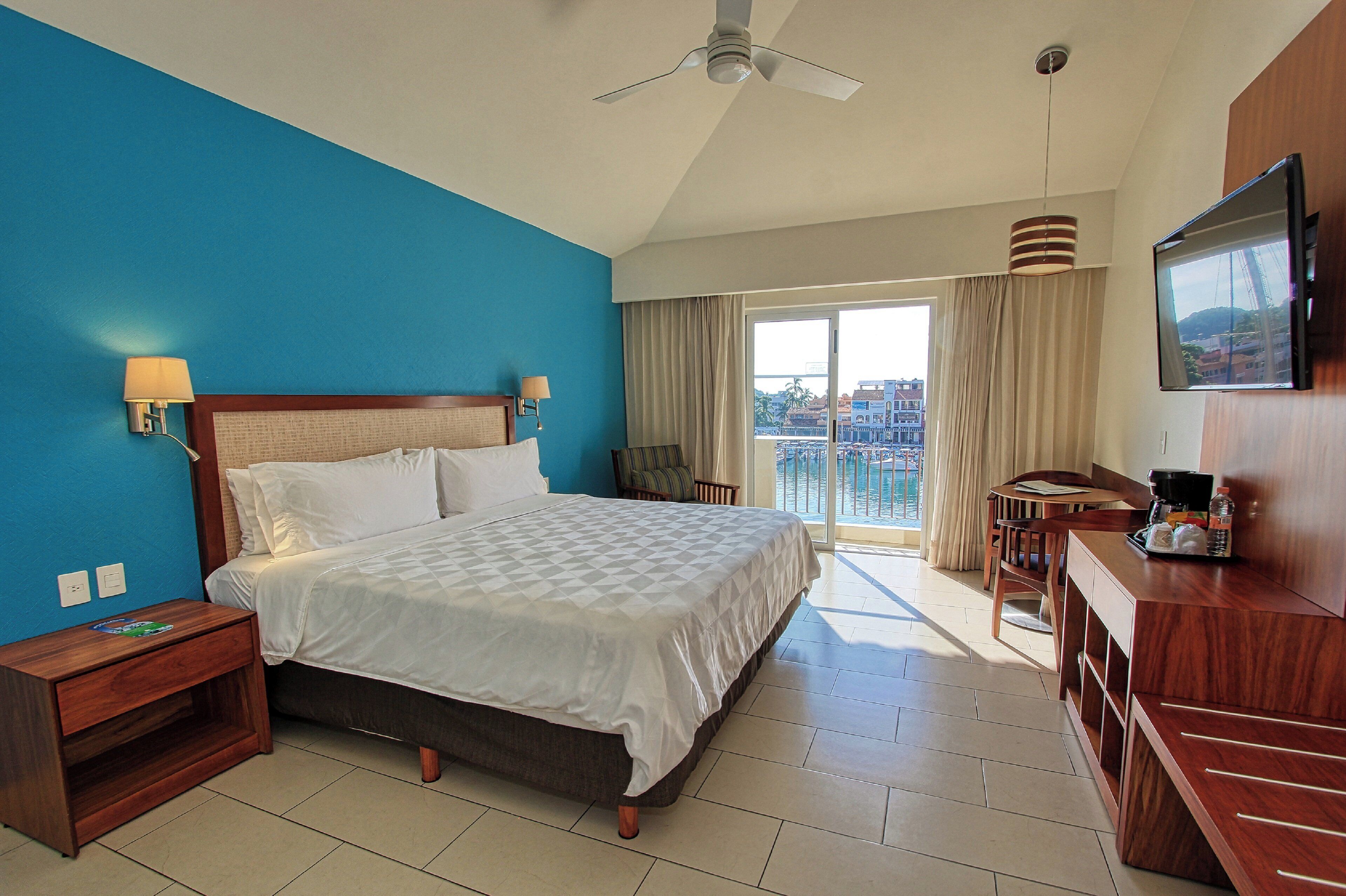 Habitación de hotel con una cama grande, un escritorio con silla, televisión y un balcón con vistas.