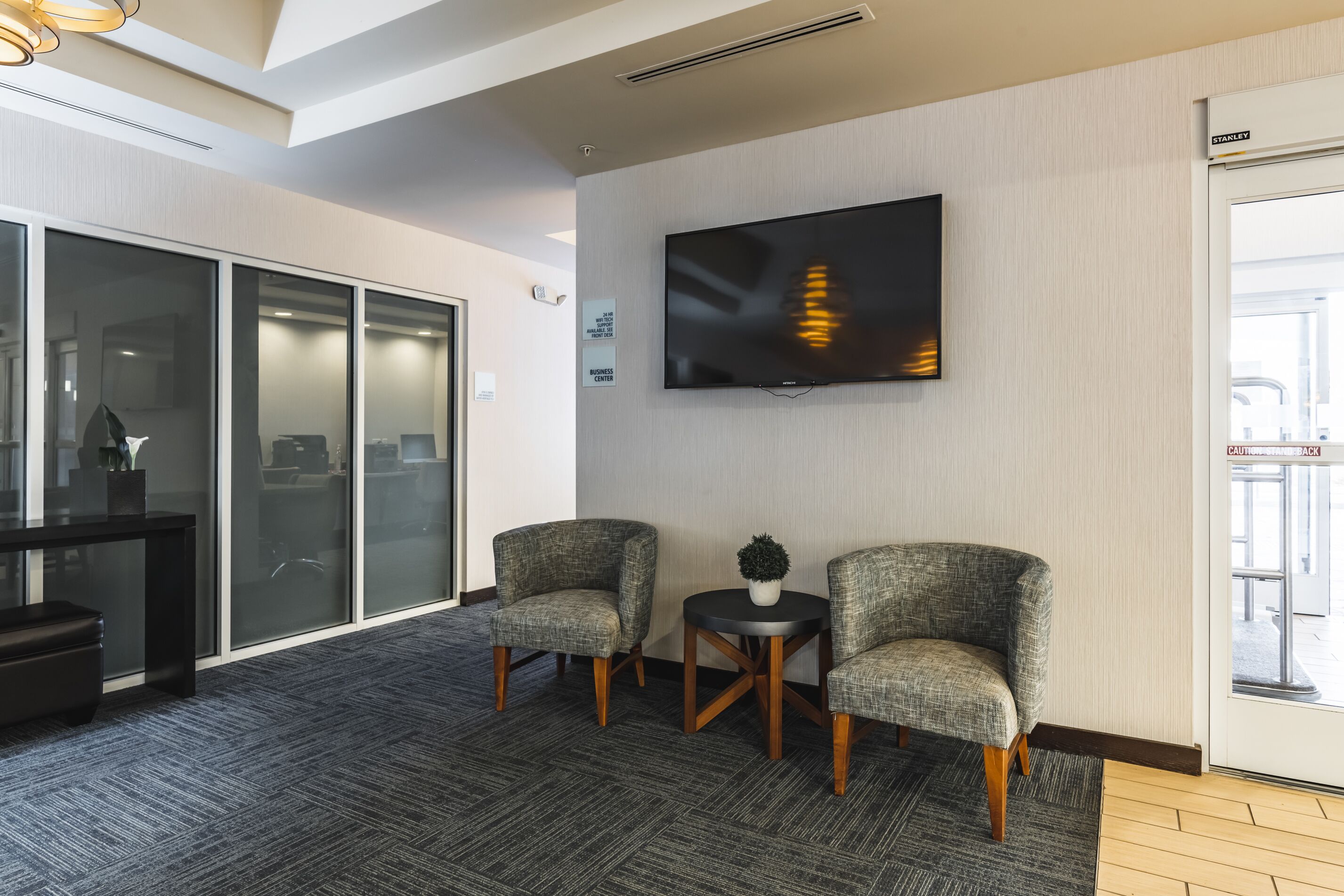 A modern waiting area with two upholstered chairs, a small table with a potted plant, a flat-screen TV mounted on the wall, and a glass door leading outside.
