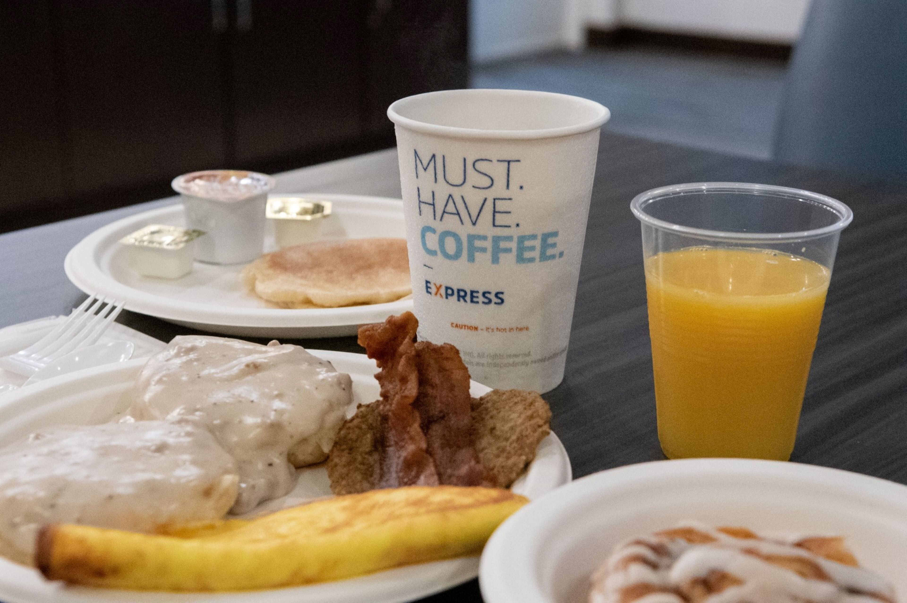 Un plato de desayuno con tortitas, tocino y una banana, una taza de café y un vaso de jugo de naranja sobre una mesa.