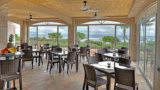 An outdoor dining area with wicker chairs and tables, large windows offering ocean views, and ceiling fans. - Hotel Tramonto (Jacó)