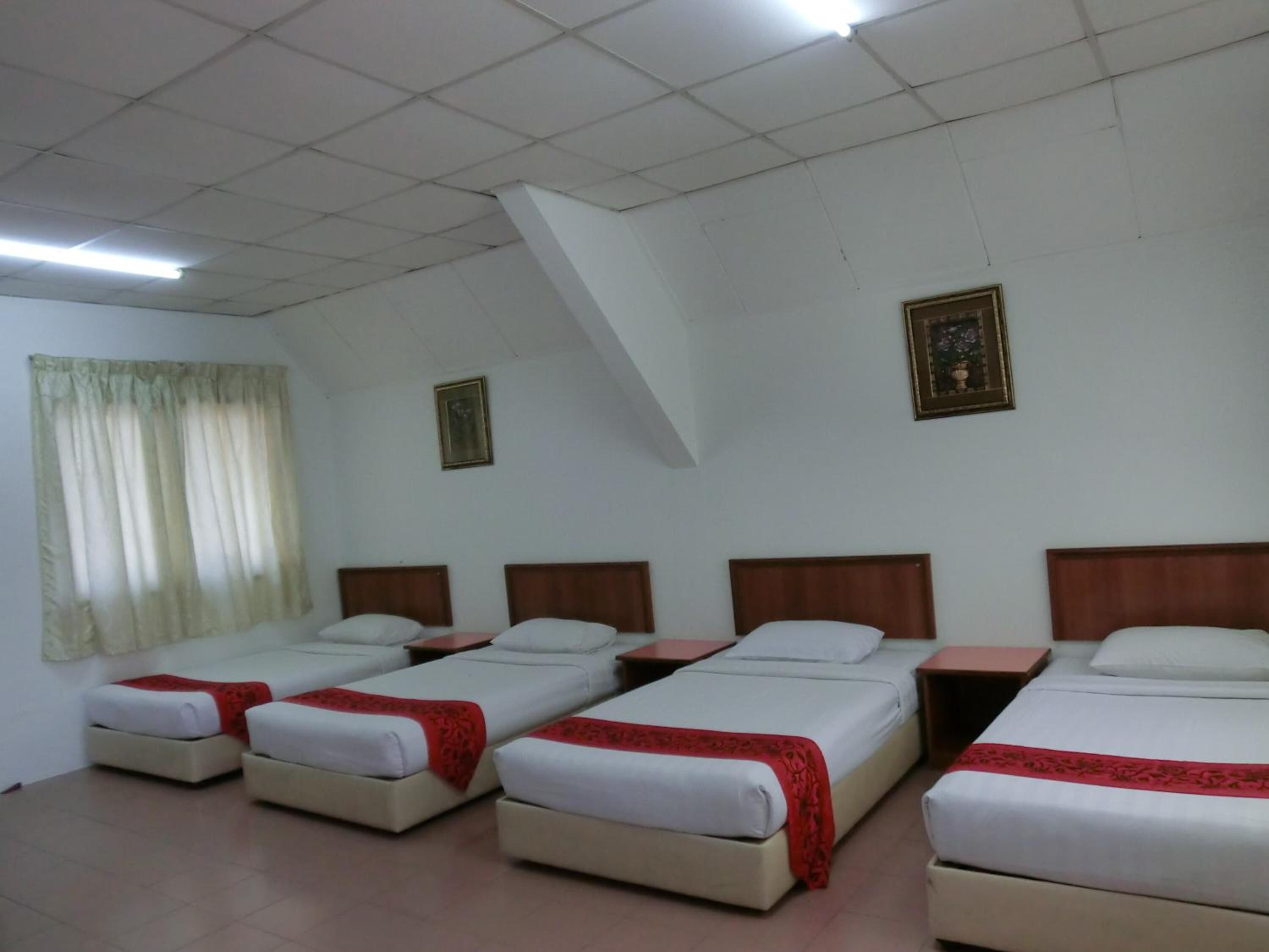 Habitació d'hotel amb quatre llits individuals, cadascun amb un capçal de fusta i roba de llit blanca. Hi ha dos quadres amb marc a les parets, una finestra amb cortines i un terra de rajola.