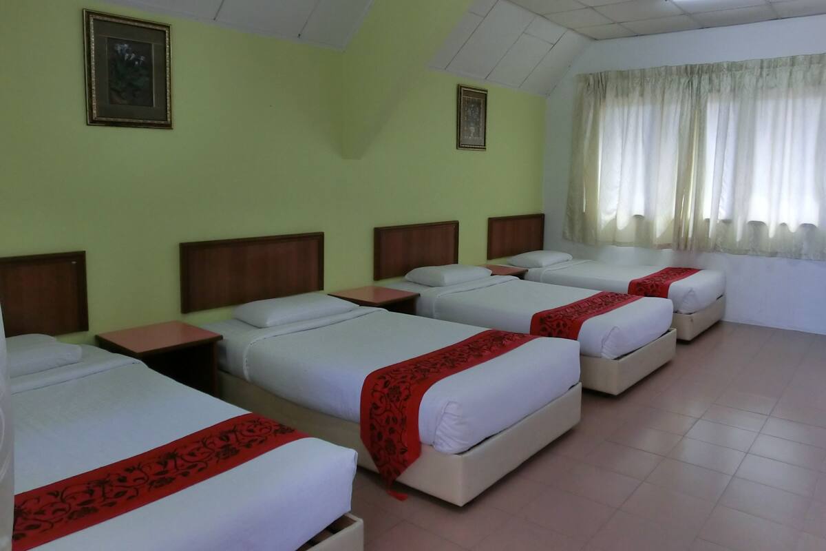 Een hotelkamer met vier eenpersoonsbedden, elk voorzien van een houten hoofdbord en een rode decoratieve kussen. De kamer heeft lichtgroene wanden, een betegelde vloer en een plafond met fluorescent verlichting.