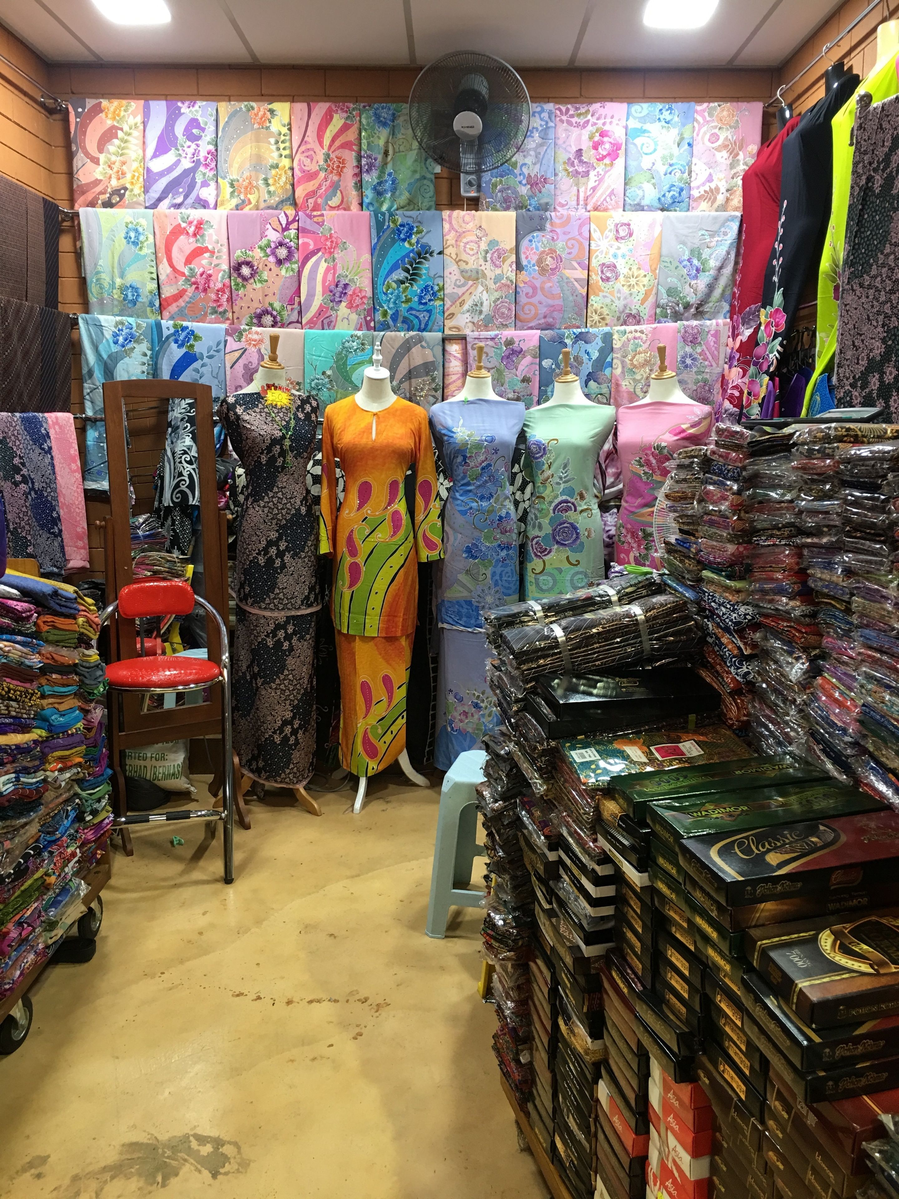 A fabric store with colorful fabrics displayed on walls and shelves, mannequins in dresses, and stacks of packaged goods.