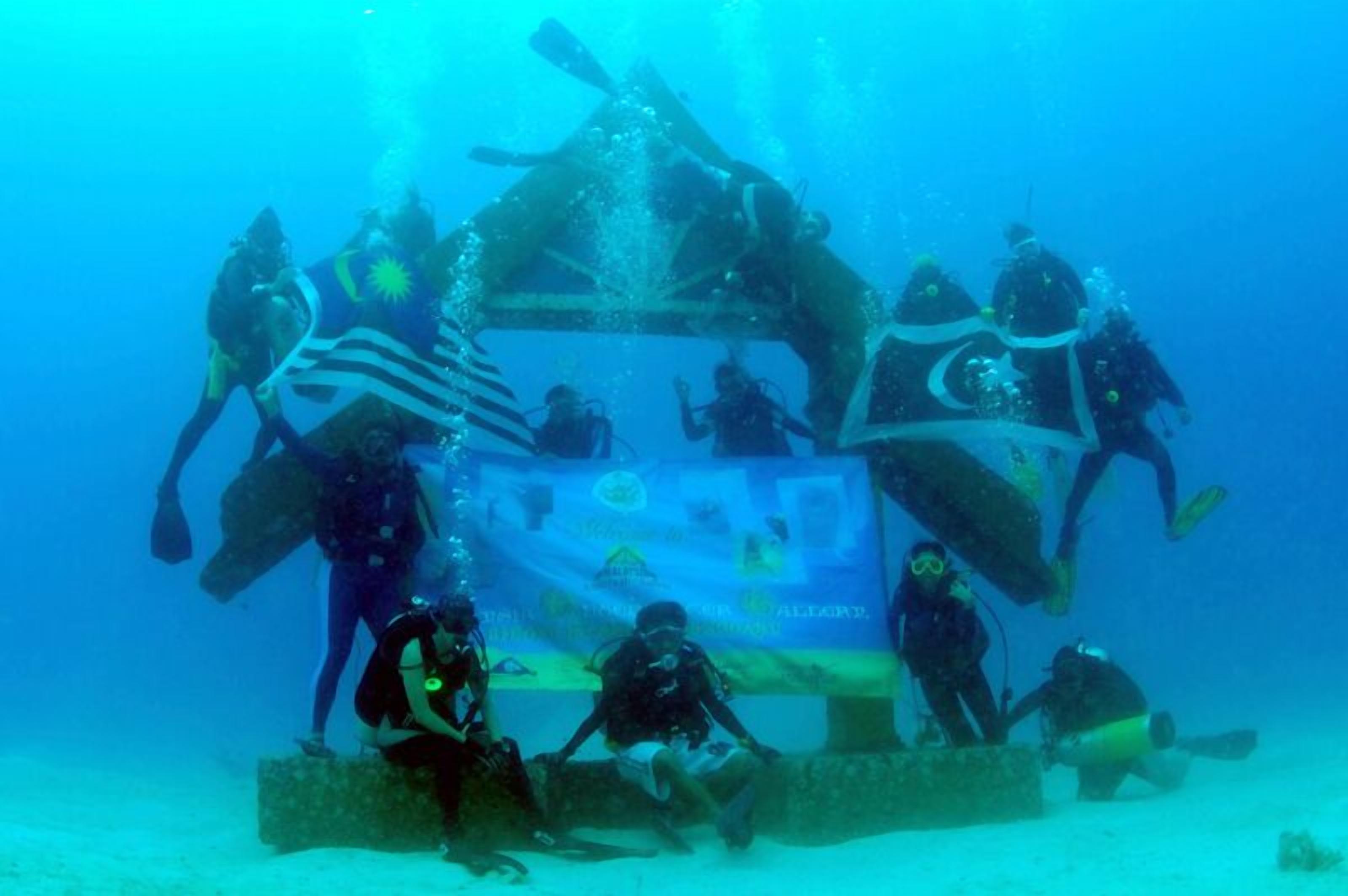 Un grupo de buzos bajo el agua, algunos de pie y otros sentados, con un cartel de fondo.
