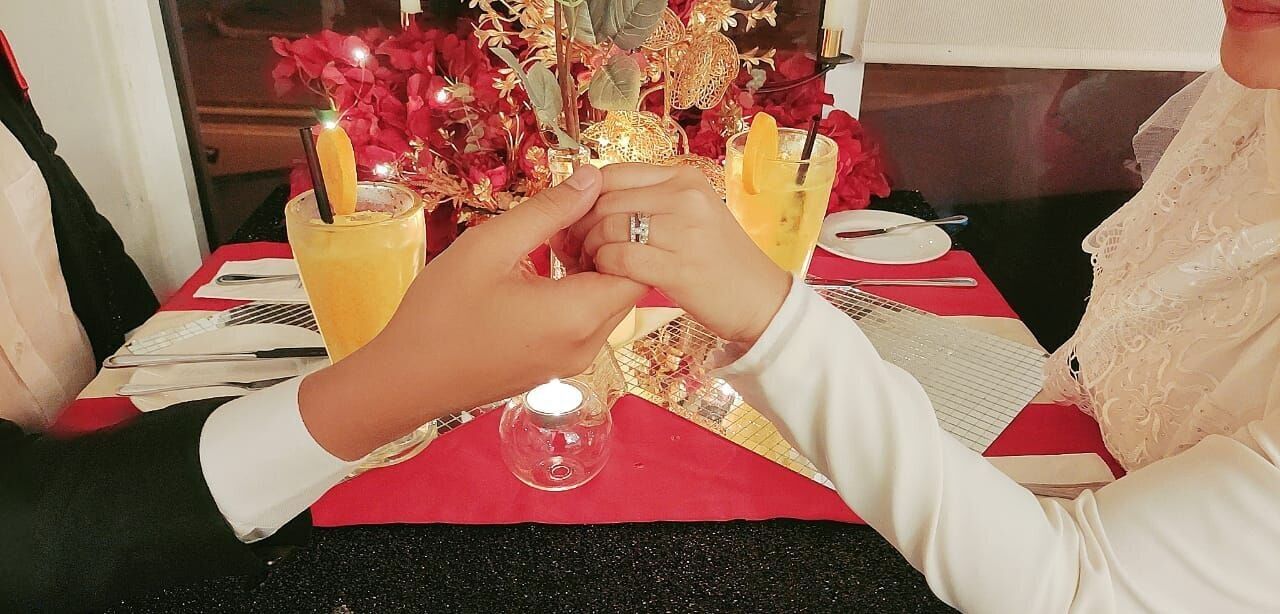 Une personne est assise à une table recouverte d’une nappe rouge. Devant elle, on aperçoit un verre contenant une boisson jaune et une petite bougie.