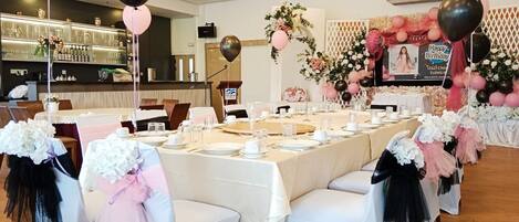 A banquet hall with round tables set for a formal event, decorated with floral arrangements and balloons.