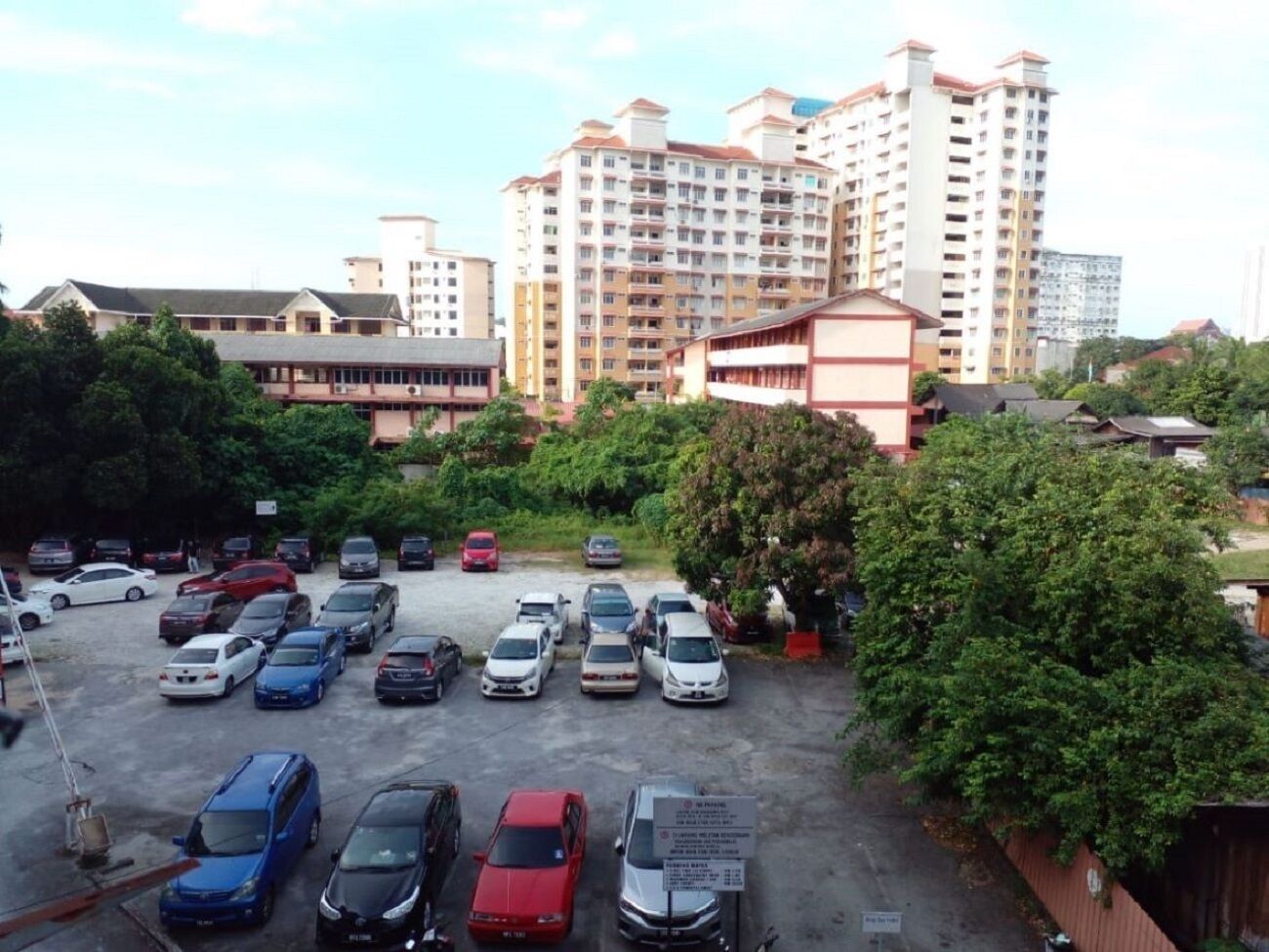 A parking lot with multiple cars, surrounded by greenery and buildings in the background.
