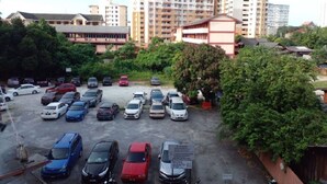 Un estacionamiento con varios automóviles, rodeado de follaje y edificios al fondo.
