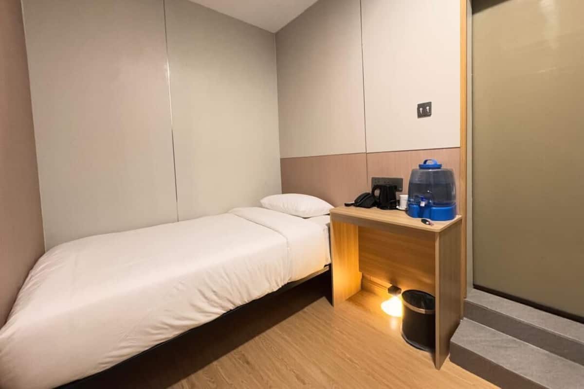 A small, modern hotel room with a single bed, a wooden desk with a coffee maker, and a trash bin.