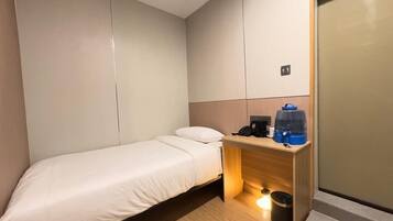 Een kleine, moderne hotelkamer met een enkel bed, een houten bureau met koffiezetapparaat en een prullenbak.