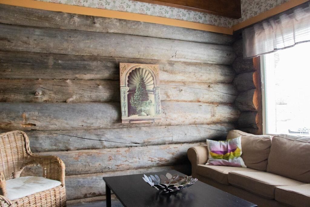 A log cabin interior with a wicker chair, a sofa, a table, and a framed picture on the wall.