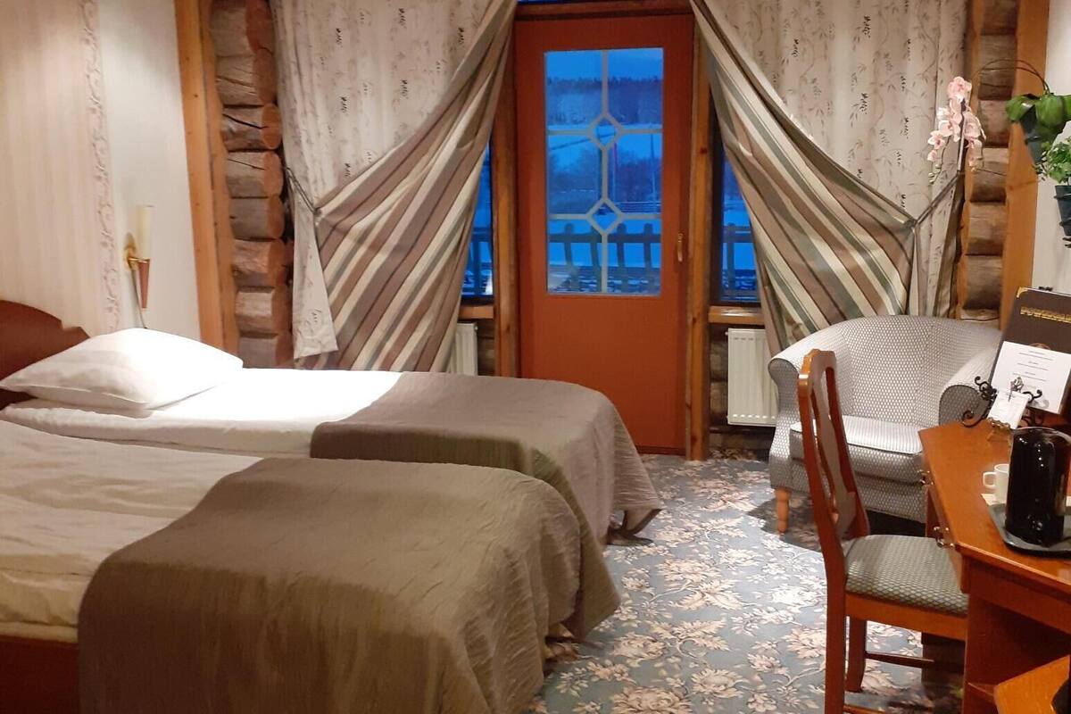 Une chambre confortable avec deux lits, une table en bois avec une bouilloire, et une vue sur l’extérieur à travers une fenêtre ornée de rideaux.