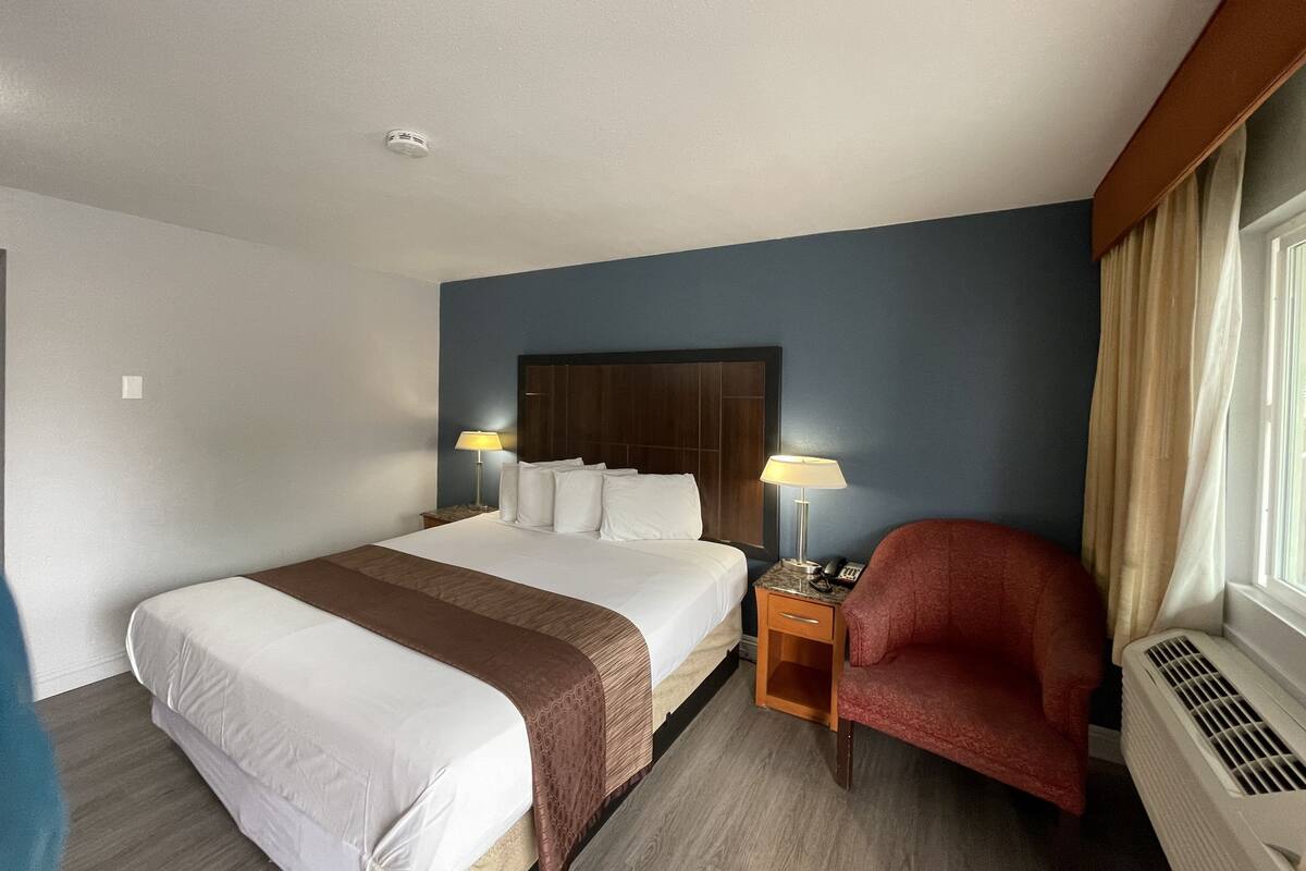A hotel room with a large bed, a wooden nightstand with a lamp, a red chair, and a window with curtains.