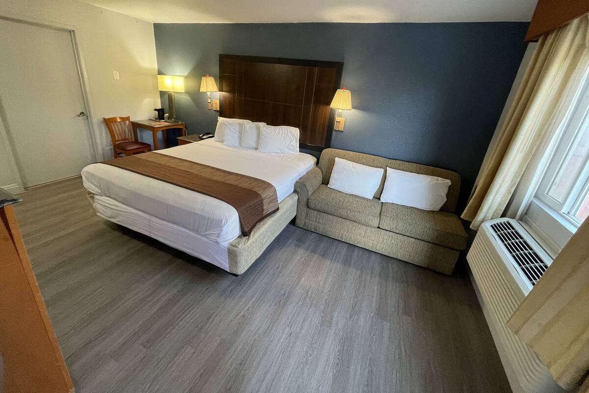 A hotel room with a large bed, a sofa, two bedside lamps, a wooden headboard, and a window with curtains.