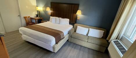 Habitación de hotel con una cama grande, un sofá, dos lámparas de noche, un cabecero de madera y una ventana con cortinas.