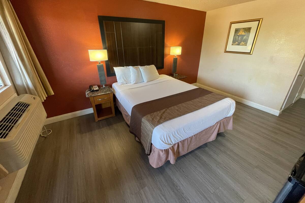 A hotel room with a large bed, two bedside lamps, a nightstand, a window with curtains, a wall-mounted air conditioner, and a framed picture on the wall.