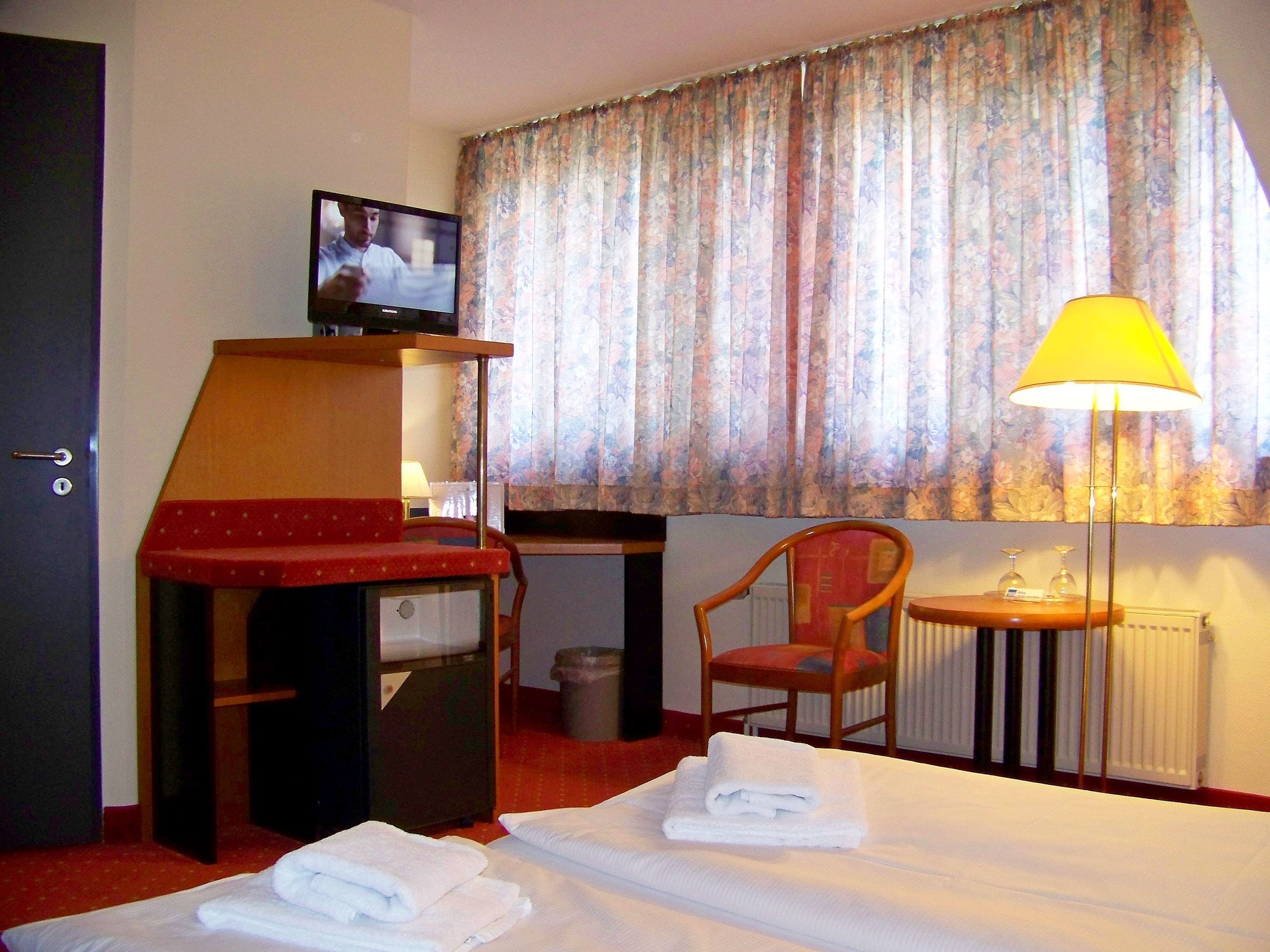 A hotel room with a bed, a desk with a television, a chair, a lamp, and a window with curtains.