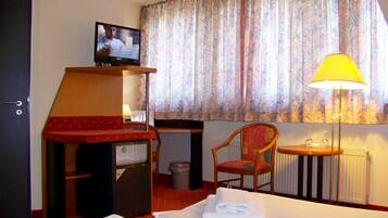 A hotel room with a bed, a desk with a television, a chair, a lamp, and a window with curtains.