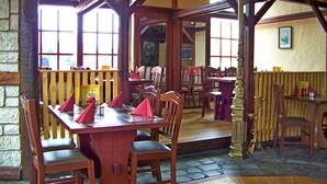 Un área de comedor al aire libre con mesas y sillas de madera, piso de piedra y ventanas amplias.