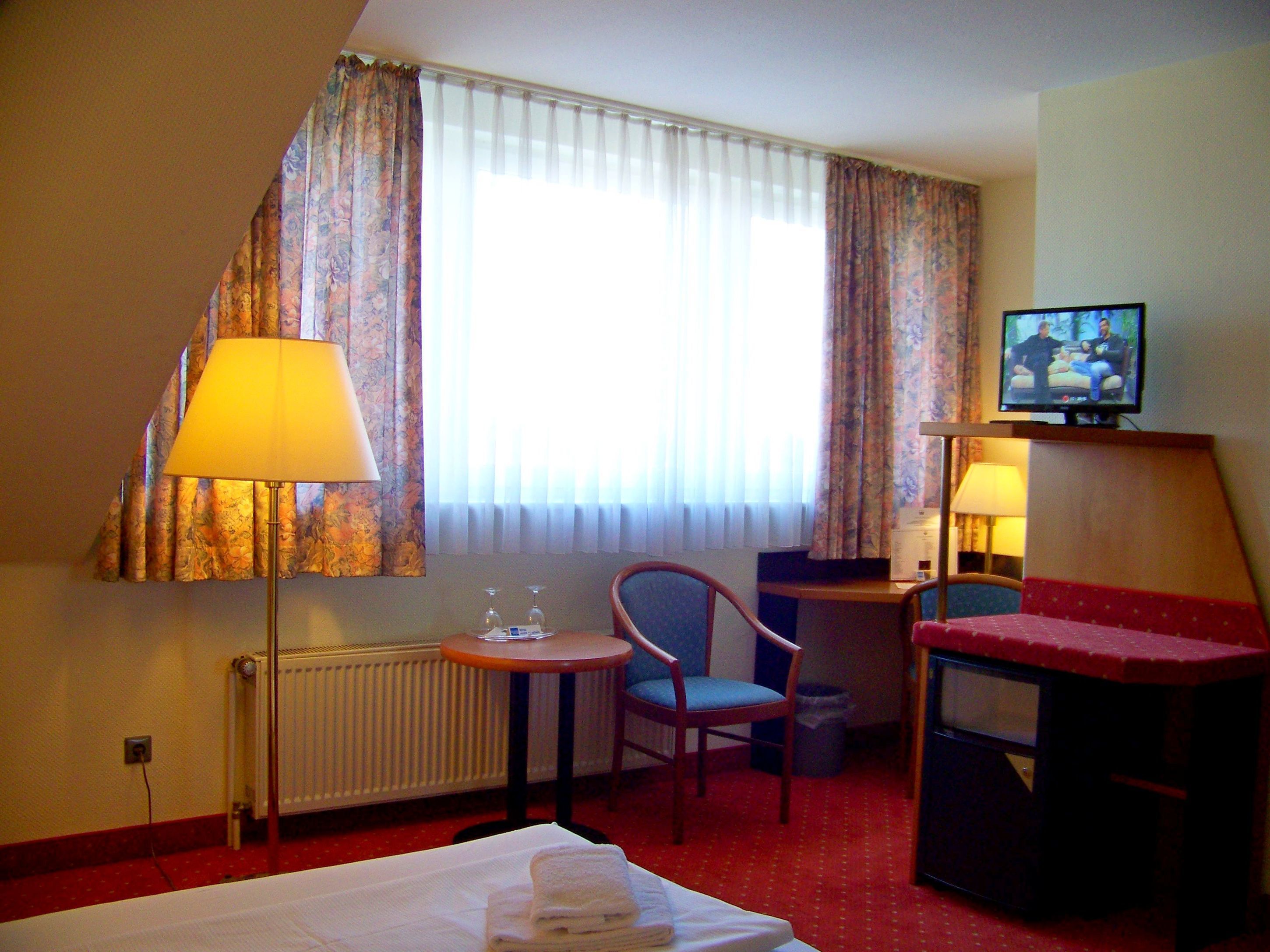 Camera d'albergo con un letto, una sedia, un tavolino, una televisione e una finestra con tende.