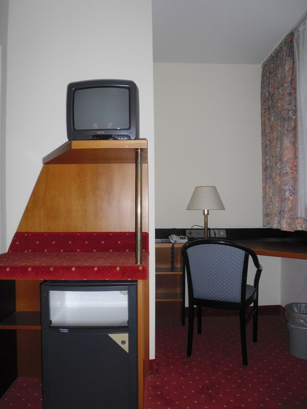 Habitación de hotel compacta con kitchenette, un pequeño televisor, un escritorio con lámpara, una silla y un cesto de basura.