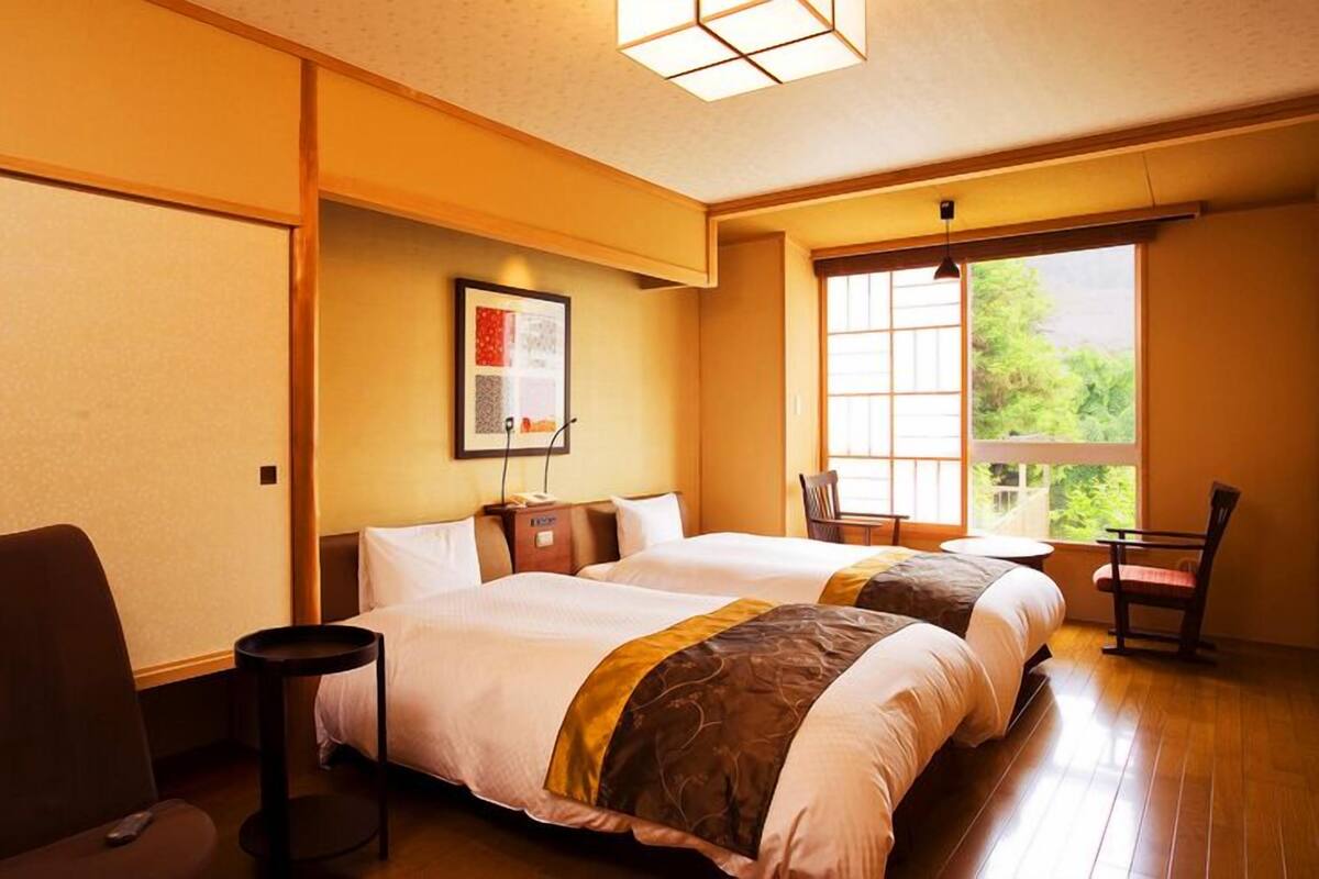 A hotel room with two beds, a small table, a chair, and a window with a view.