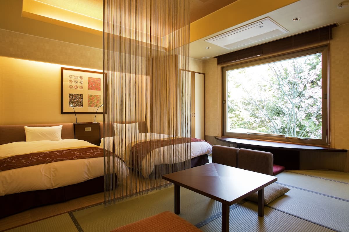 A hotel room with a bed, a small table, and a window with a view of trees.