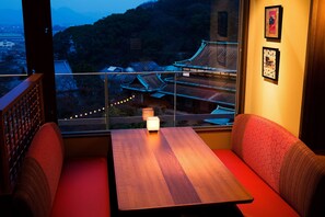 A room with a wooden table and red cushioned seating, a window with a view of traditional buildings, and framed pictures on the wall.
