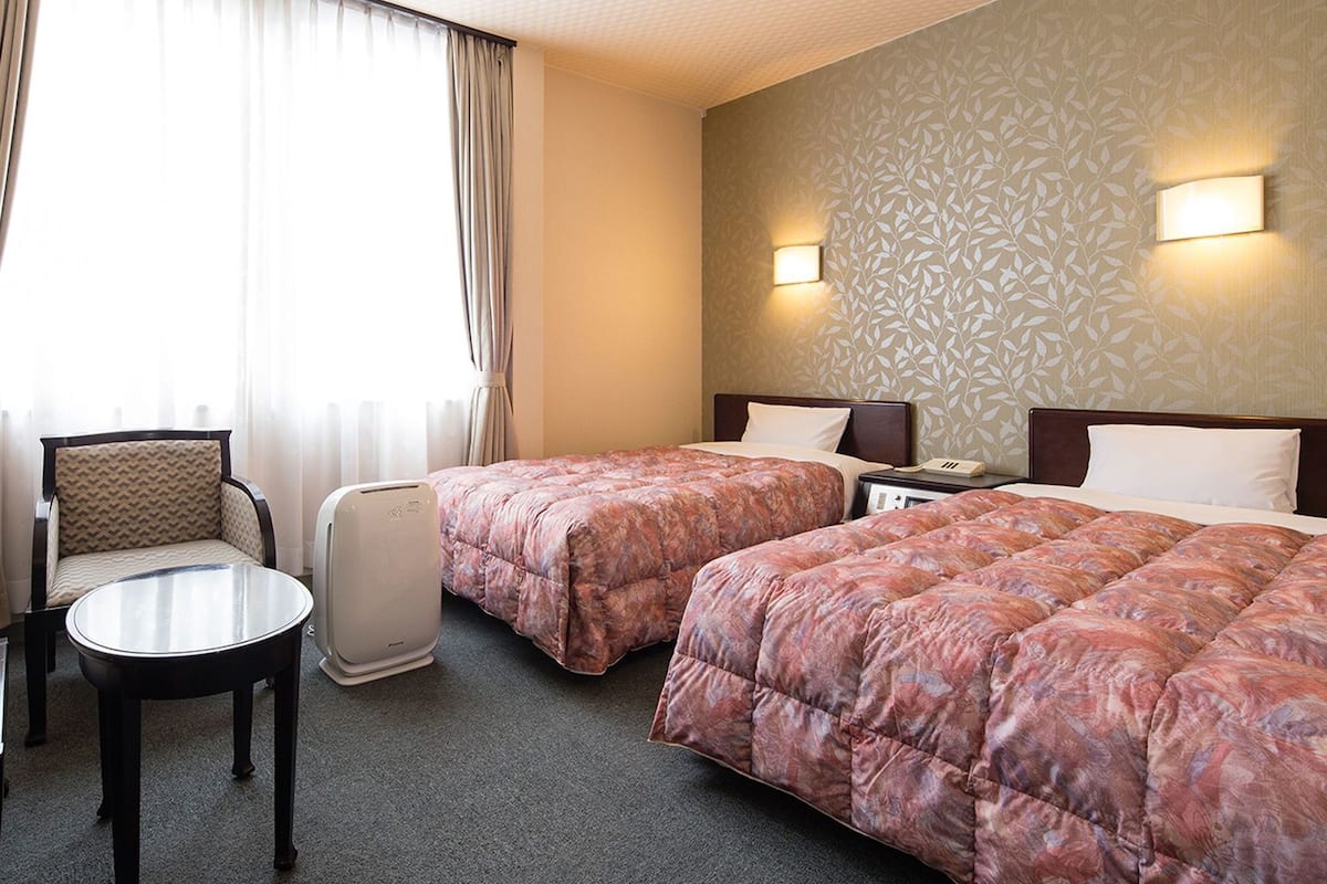A hotel room with two beds, a chair, a table, and a wall-mounted phone.