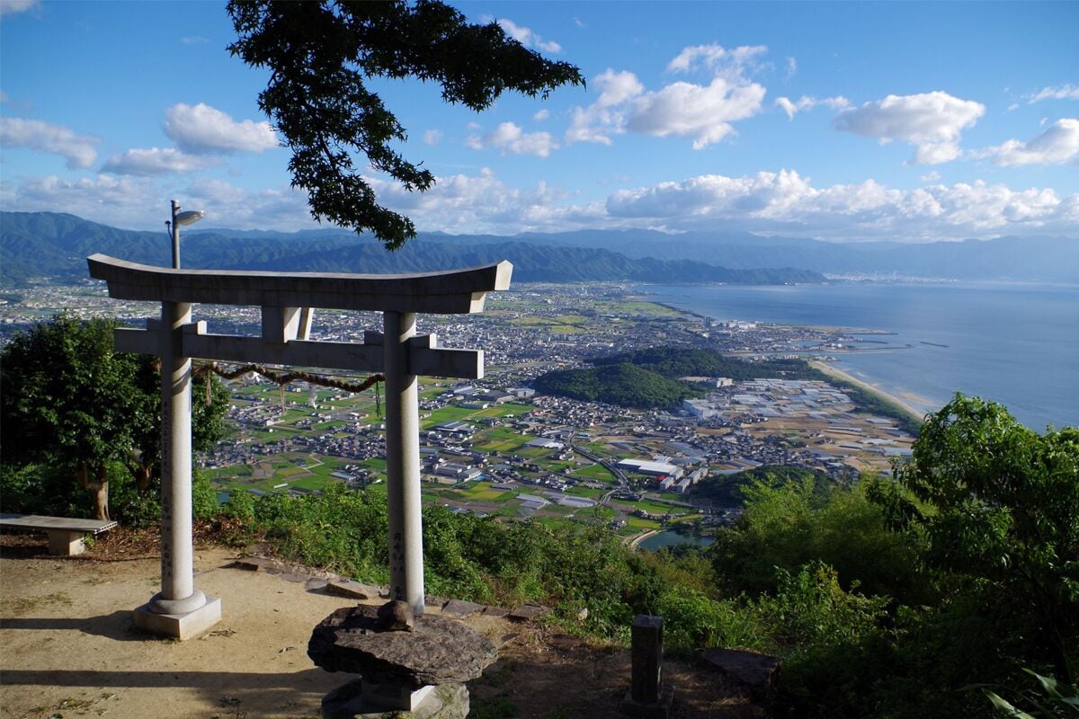 Perinteinen japanilainen torii-portti, josta avautuu näkymä rannikkokaupunkiin, ja taustalla kohoavat vuoret.
