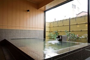 A hot spring bath with a view of a bamboo fence and greenery outside. - Kotohira River Side Hotel (Kotohira)