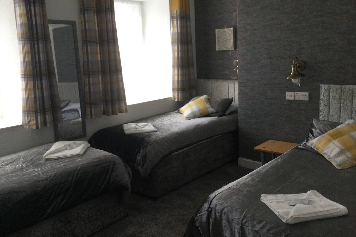 A room with two beds, a small bedside table, a wall-mounted light, and a window with curtains.