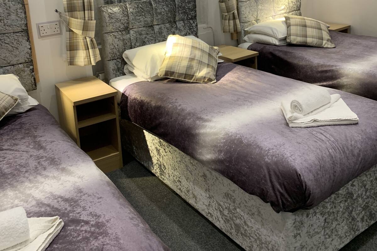 A hotel room with three beds, a large window with patterned curtains, a stone-textured headboard, and a wooden hanger on the wall.