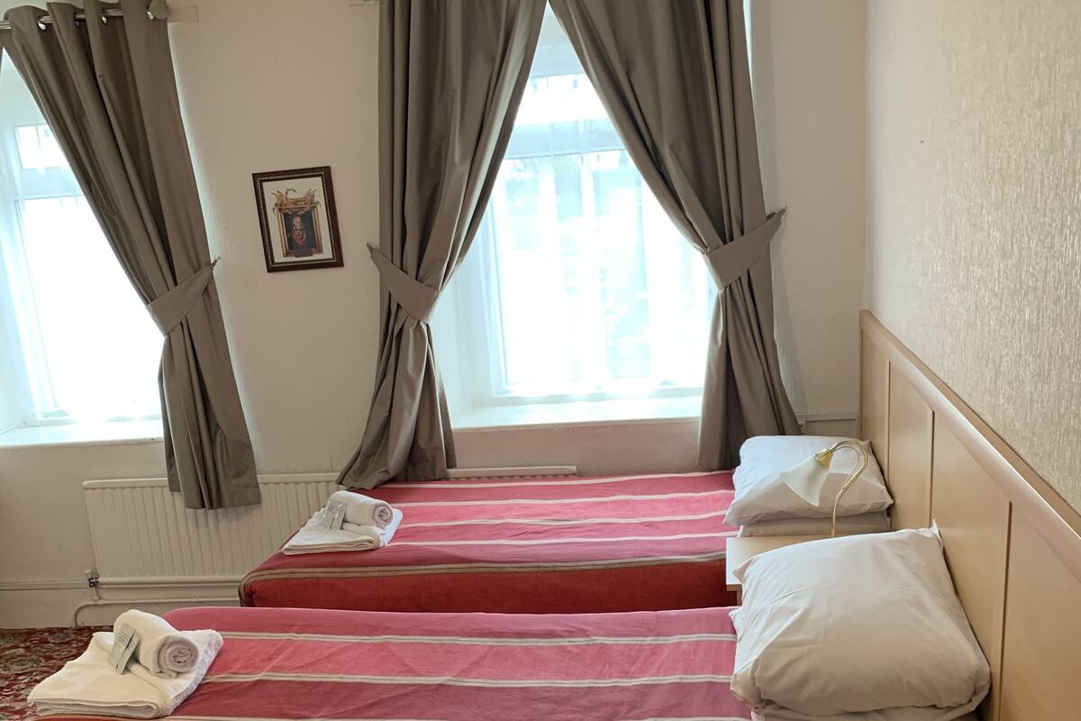 A hotel room with two beds, each with a red-striped bedspread, white pillows, and a folded towel. There is a wooden nightstand with a lamp, and a radiator on the wall.