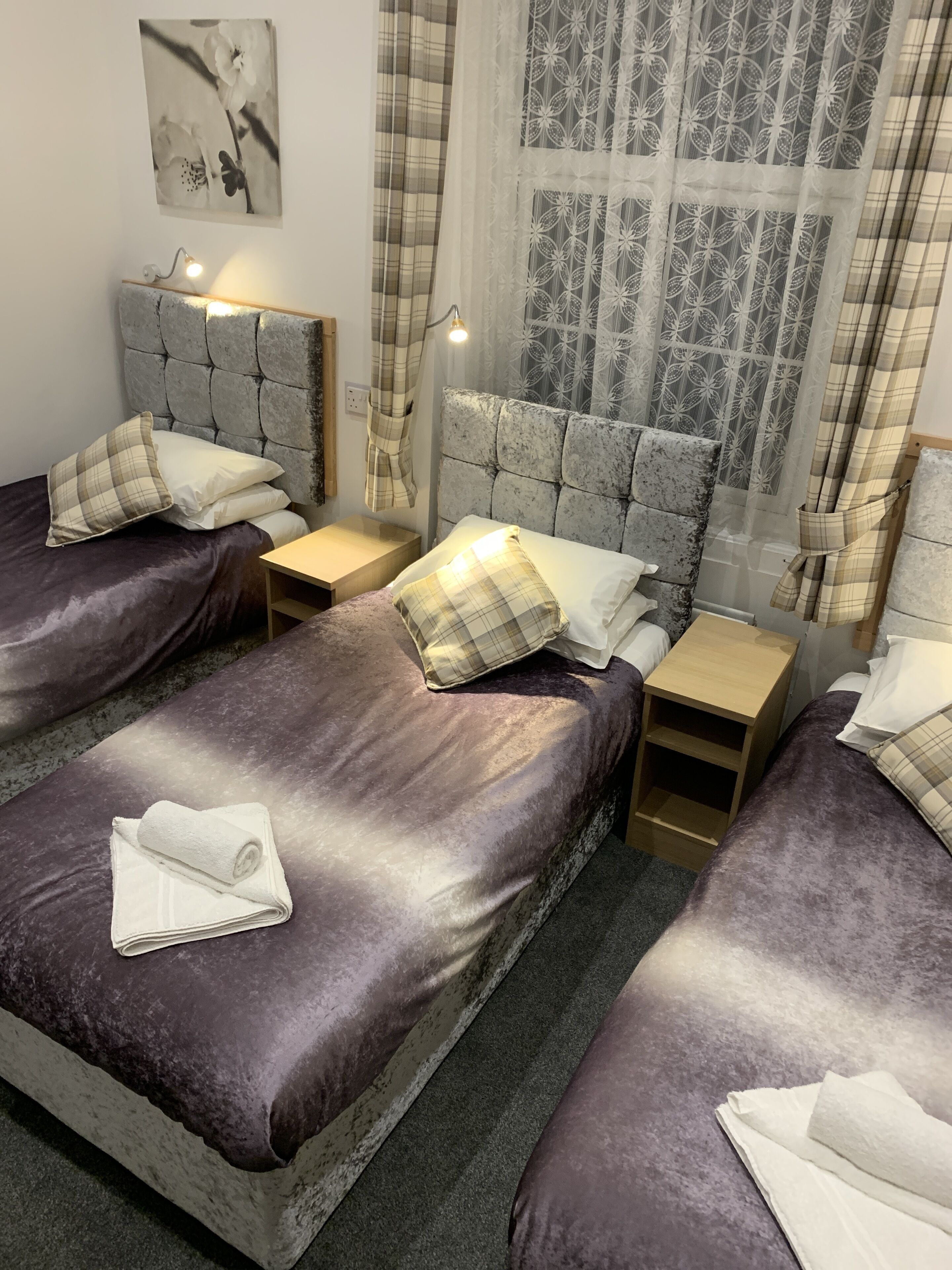 A hotel room with two beds, each with a headboard and a nightstand. There is a window with patterned curtains, a wall-mounted light, and a framed picture on the wall.