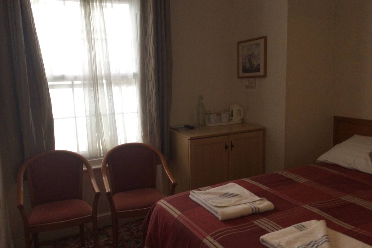 A hotel room with a bed, two chairs, a small table with a kettle, and a window with curtains.